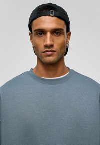 Overmaatse blauwe sweatshirt met een gestructureerde afwerking, ronde hals en subtiele ribbels, gecombineerd met een zwarte baseballpet.