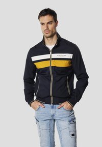 Námořnická bunda s žlutými a bílými vodorovnými pruhy, přiléhavý střih, zapínání na zip a elastické manžety. Nošená přes bílou košili a doplněná světlými džíny.