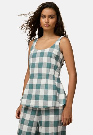 Žena s dlhými tmavými vlasmi má na sebe zelený a biely károvaný top bez rukávov a k nemu ladné široké nohavice na jednofarebnom pozadí.