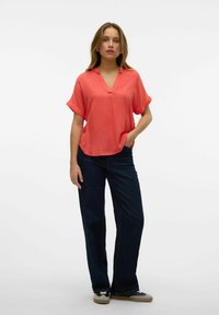 Blusa corallo a maniche corte con scollo a V e tasca; abbinata a jeans svasati scuri e sneakers grigie con una accentuazione bianca.