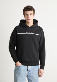 Črna kapuca s sivim logotipom "Calvin Klein" in horizontalno črto. Ima sproščen kroj in mehak material. Oblečena je čez svetlo modre kavbojke.