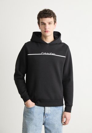 Čierna mikina s kapucňou, na ktorej je biele logo "Calvin Klein" a horizontálny pruh. Vyznačuje sa uvoľneným strihom a mäkkou textúrou. Nosená cez svetlomodré džínsy.