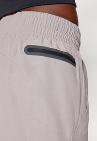 Närbild på en person som bär ljusgrå byxor med elastisk midjeresår och en svart blixtlåsficka på höger sida.