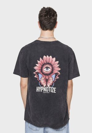 Una camiseta de algodón negra presenta un gran gráfico de un girasol, un ojo y mariposas, con la palabra "HIPNOTIZAR" en una fuente en negrita en la parte inferior.