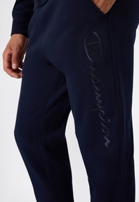 Marineblauwe joggingsbroek gemaakt van zachte stof met een verticale geborduurde "Champion" logo op het linkerpijp en zijzakken.