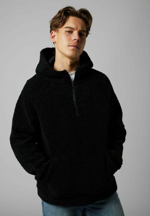 Ung mand med lysebrunt hår iført en sort sherpa fleece-hættetrøje og blå jeans, hænderne i lommerne, neutral grå baggrund.