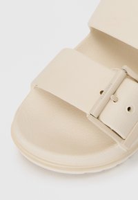Sandalo scivoloso di un beige chiaro realizzato in materiale sintetico testurizzato. Caratterizzato da una fascia ampia con una fibbia rettangolare e una suola arrotondata.