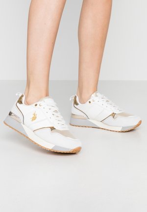 Zapatillas blancas con detalles dorados y suelas beige usadas en los pies contra un fondo liso gris claro.