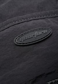 Black fabric with a textured surface; features a raised oval rubber logo applied along a stitched seam.