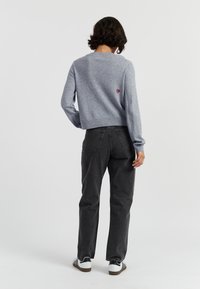 Een persoon met kort, krullend haar draagt een grijze trui en donkere spijkerbroeken, met de rug naar ons toe gekeerd. Zwart-witte sneakers maken de outfit compleet.