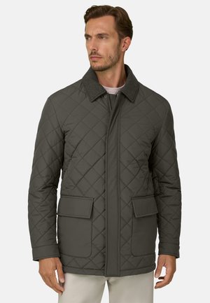 Mann trägt eine dunkelgrüne gesteppte Jacke mit großen Fronttaschen und einem kontrastierenden Kragen, schaut seitlich vor einem einfarbigen Hintergrund.