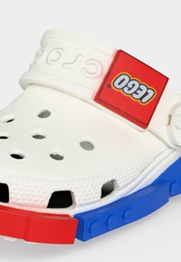 Λευκό clog Crocs με οπές αερισμού, κόκκινο λογότυπο LEGO στο λουράκι και κόκκινα και μπλε διακοσμητικά στοιχεία στο πέλμα με καρφιά.