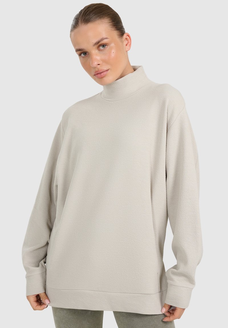 Jeune femme aux cheveux tirés en arrière, portant un pull ample à col roulé beige clair et un pantalon gris clair, debout devant un fond uni.