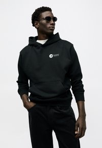 Άνδρας με μαύρο φούτερ με κουκούλα και λογότυπο New Balance, γυαλιά ηλίου και μαύρο παντελόνι, στέκεται με το ένα χέρι στην τσέπη μπροστά σε λευκό φόντο.