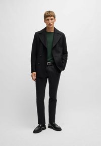 Cappotto doppio petto nero sopra una camicia verde scuro, abbinato a pantaloni e scarpe neri. Il cappotto presenta ampi revers e bottoni.