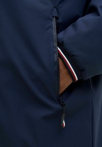 Marineblaue Jacke mit einer seitlichen Reißverschlusstasche und einem gerippten Bündchen, das in Rot, Weiß und Blau gestreift ist. Glatte Stofftextur.