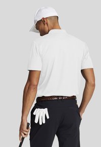 Uomo che indossa un cappello bianco e una polo, pantaloni neri con guanti da golf bianchi riposti nella tasca posteriore, mentre tiene un bastone da golf e guarda verso l'esterno.