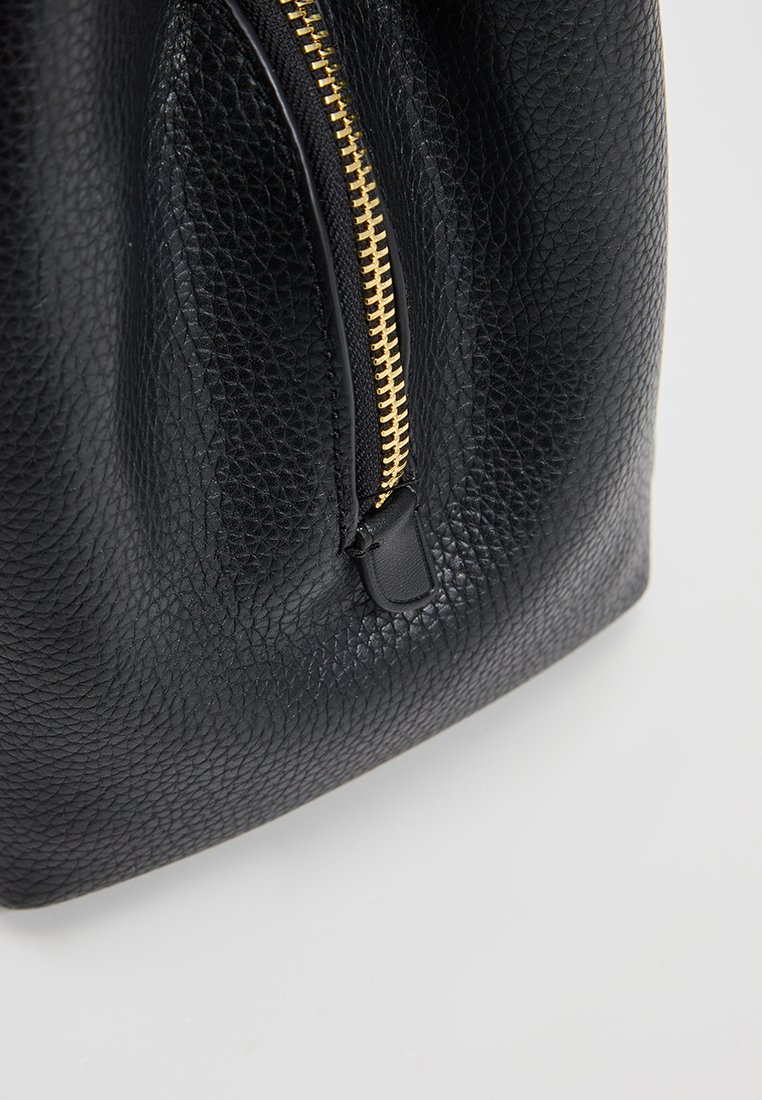 Zwarte leren tas met textuur, voorzien van een gouden rits, met een ronde vorm en een zachte structuur. De rits heeft een kleine leren accent.