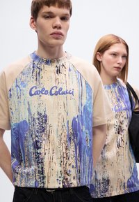 Μπλούζα με κοντά μανίκια, μπεζ βάση και μπλε αφηρημένο σχέδιο. Έχει στρογγυλή λαιμόκοψη και λογότυπο σε μοβ χρώμα στο στήθος.