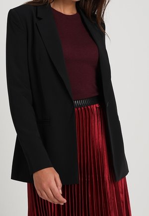 Femme portant un blazer noir sur un top bordeaux foncé et une jupe plissée rouge brillante, main posée à côté.