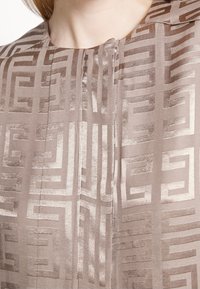 Blusa in seta di un leggero beige con un evidente motivo geometrico in una tonalità più scura. Presenta un colletto e dettagli con pieghe frontali.