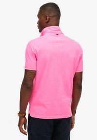 Polo à manches courtes en coton rose fluo, doté d'un col montant, d'un détail avec boutons dans le dos, et d'une coupe décontractée. Bras dégagé avec un short foncé.