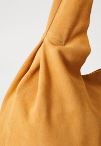 Tan suede shoulder bag with a soft texture, featuring gathered detailing at the shoulder strap and neat stitching along the seams.