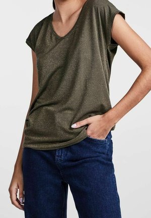 Vrouw draagt een olijfgroen gestreept T-shirt met korte mouwen en donkerblauwe spijkerbroek, met één hand in de zak en een neutrale achtergrond.