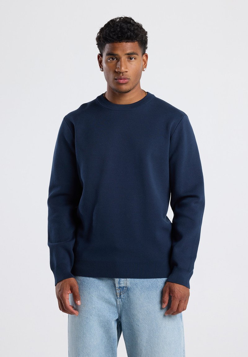 Tummansininen collegemekko, jossa on pyöreä kaula-aukko ja pitkät hihat. Varusteena ribbineuloksesta valmistettu helma ja hihansuut, valmistettu sileästä ja pehmeästä kankaasta.