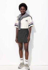 Jeune femme aux cheveux courts et bouclés portant une chemise crème avec le chiffre "75", une jupe noire, un pull gris drapé sur les épaules, des chaussettes blanches et des baskets.