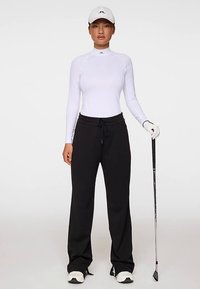 Vit långärmad topp, svart löst sittande byxor, vit keps, håller i en golfklubba med svarta detaljer. Kläderna är i slät, andningsbar tyg.