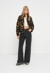 Joven mujer de pie con una mano en el bolsillo, vistiendo una chaqueta de patrón negro y marrón, una camiseta blanca de corte crop, pantalones negros de pierna ancha y zapatillas grises.