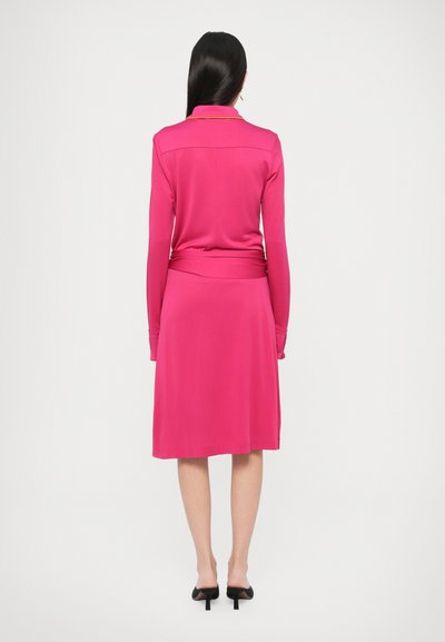 Różowa sukienka z długim rękawem, z kołnierzykiem, dopasowaną talią i rozkloszowanym dołem. Tkanina wydaje się gładka i lekko elastyczna.