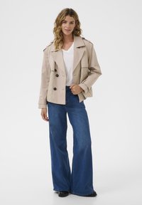Kvinde med bølget hår iført en beige quiltet jakke, hvid v-hals skjorte, brede blå jeans og sorte sko, stående mod en hvid baggrund.