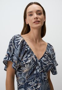 Marine jurk met bloemmotief, korte fladdermouwen en een V-halslijn. Zachte stof met een fijne glans en gathered details bij de buste.