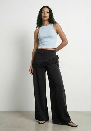 Femme aux cheveux bouclés portant un crop top sans manches bleu clair, un pantalon large noir avec un détail en dentelle, et des sandales noires, debout contre un mur uni.