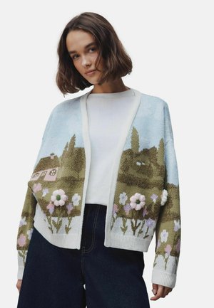 Young woman wearing a cardigan with a countryside landscape and 3D floral details over a white top and dark pants.