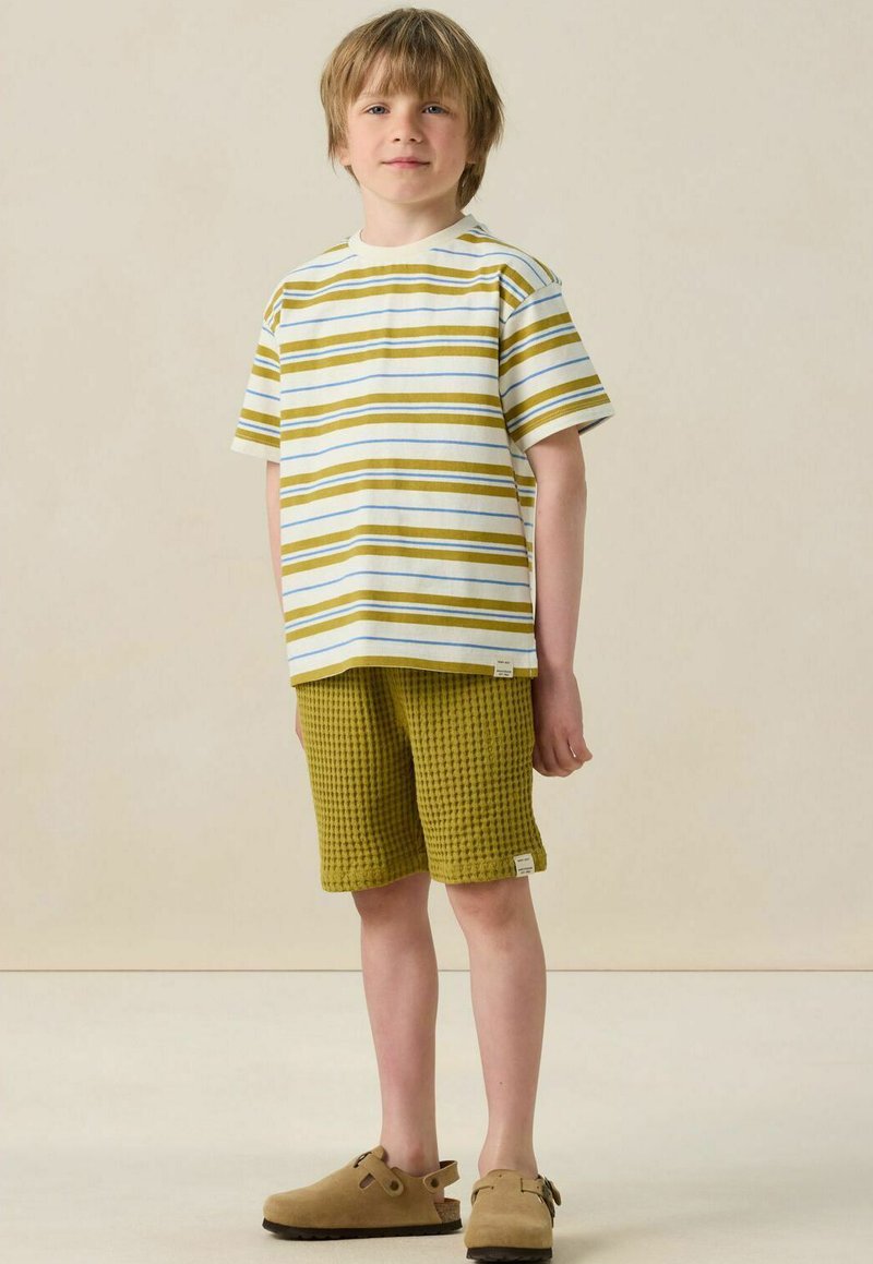 Chemise à manches courtes rayée en blanc et vert olive, associée à un short en vert olive texturé. Des chaussures slip-on beiges complètent la tenue.