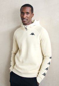 Man wearing cream hoodie with black logo on chest and sleeves, standing against a light textured wall, hands in pockets, slight smile.