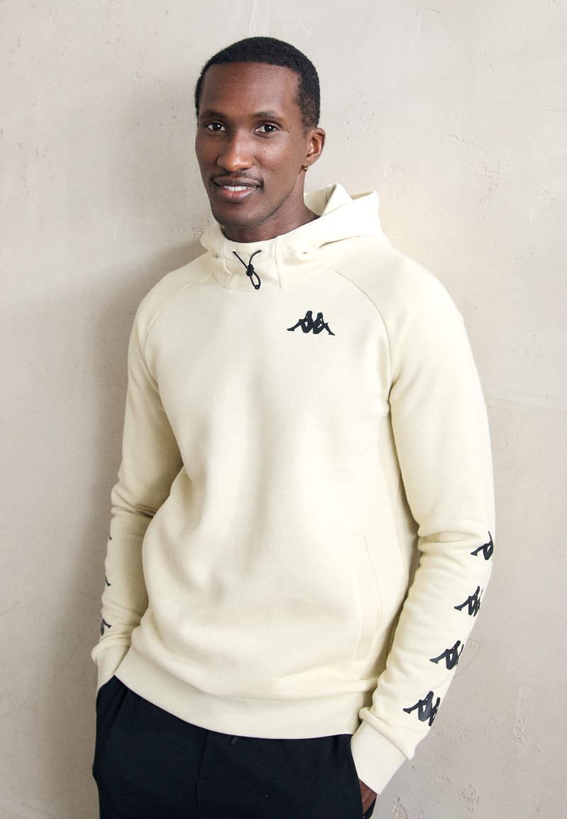 Man wearing cream hoodie with black logo on chest and sleeves, standing against a light textured wall, hands in pockets, slight smile.