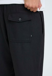 Zwarte stoffen broeken met een knoopzak en een klein label op de zijkant, met een gladde textuur en een ontspannen pasvorm.
