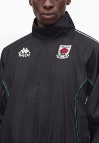 Personne portant une veste Kappa noire avec un blason en forme de rose où il est écrit "Authentic Fondato in Italia,", visible des épaules au milieu de la poitrine.