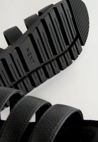 Schwarze Sandalen mit strukturierten Riemen und einer dicken, geriffelten Gummisohle. Die Laufsohle trägt das eingestanzte Wort "NEXT".