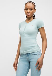 Νεαρή γυναίκα με πλεγμένα μαλλιά, φορώντας ένα ανοιχτό γαλάζιο πουκάμισο με κοντά μανίκια και κουμπιά, και ανοιχτό γαλάζιο τζιν, στέκεται μπροστά σε απλό φόντο.