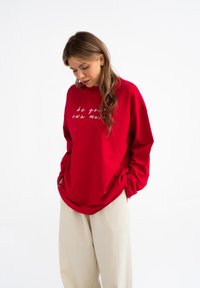 Czerwony sweter z luźnym krojem i długimi rękawami, z białym napisem z przodu. Noszony z jasnobeżowymi spodniami, prezentując casualowy styl.