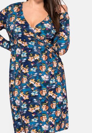 Mujer con vestido cruzado de manga larga azul con flores naranjas, blancas y rosas, de pie frente a un fondo blanco.