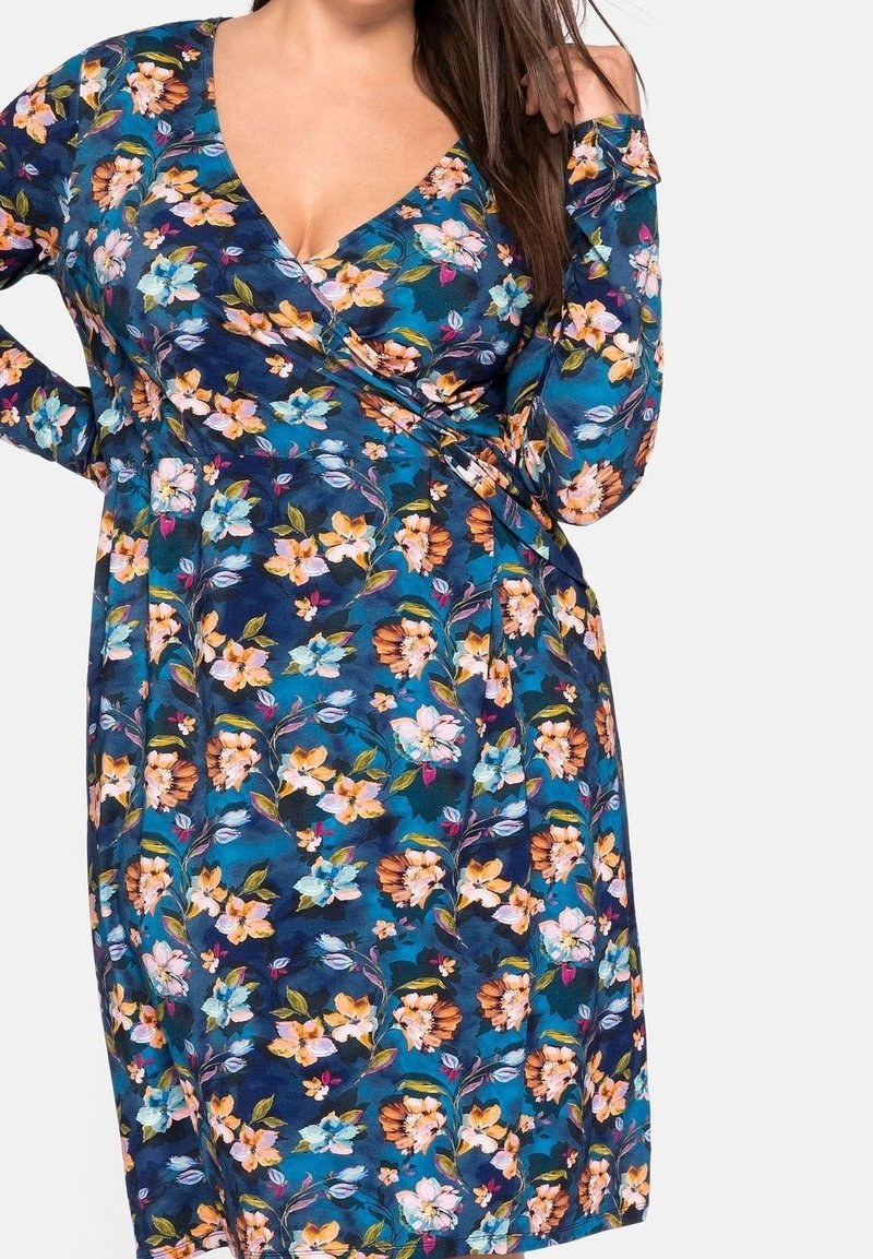 Femme portant une robe portefeuille bleue à manches longues avec des fleurs orange, blanches et roses, debout sur un fond blanc.