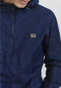 Veste légère bleu marine avec un demi-zip, capuche ajustable et un petit patch logo carré sur la poitrine. Matériau lisse et résistant à l'eau.
