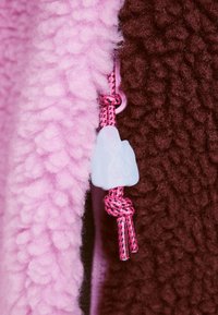 Fleecejacka med en fluffig rosa del och en brun strukturerad del, med en rosa flätad dragsko och en ljusblå dekorativ knopp.
