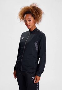 Schwarze Sportjacke mit Reißverschluss und hohem Kragen, mit grauen Akzenten und einem strukturierten Design. Kombiniert mit schwarzen Sporthosen.